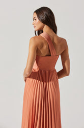 A woman is wearing a sleeveless, coral-colored dress with a pleated skirt. The top part of the dress features a textured, smocked design with a wide, single shoulder strap on one side, which extends over the shoulder and down the back. The dress's fabric appears lightweight and has a sheen that enhances the pleats.